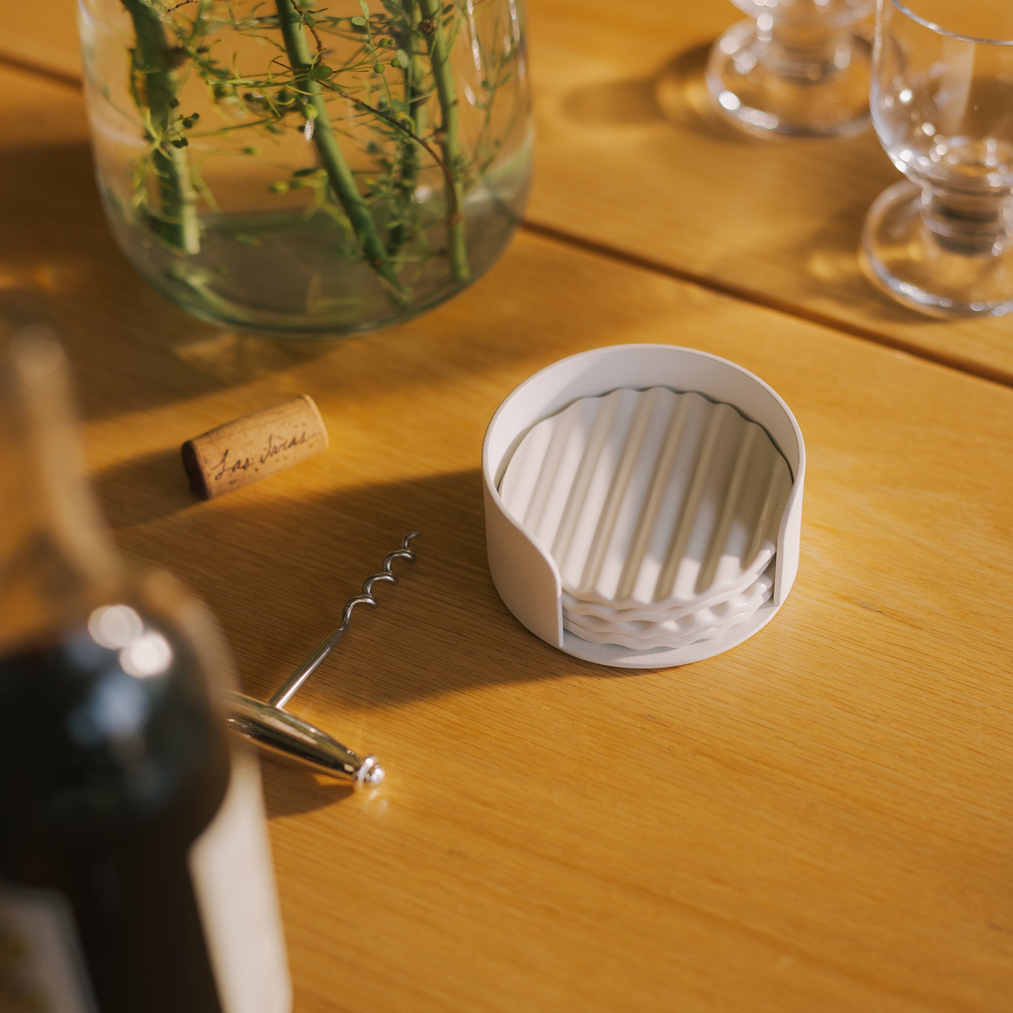 White ribbed ceramic coaster set in round holder on oak dining table beside wine cork and corkscrew