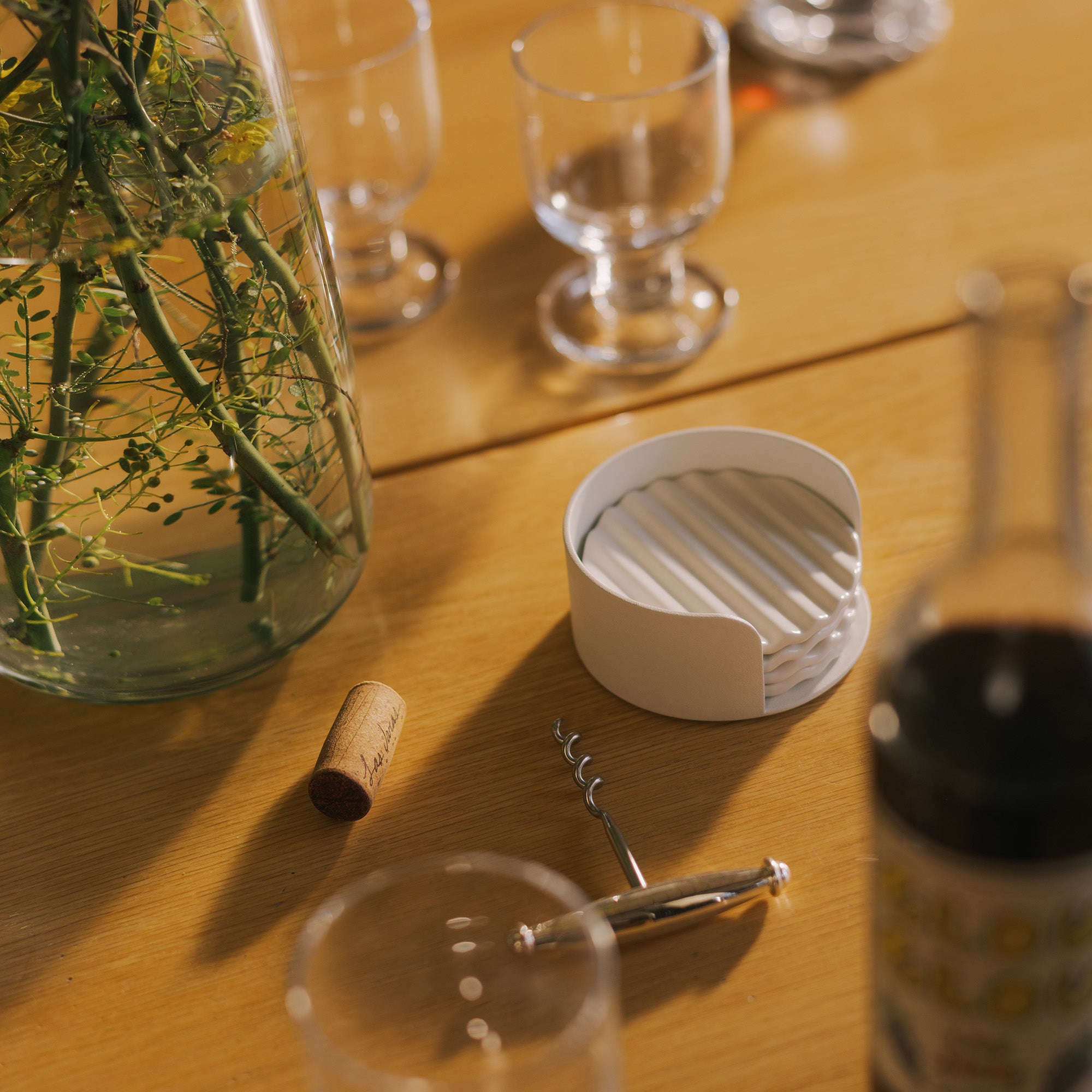 White ribbed ceramic waved coaster set in round holder on oak dining table beside wine cork and corkscrew