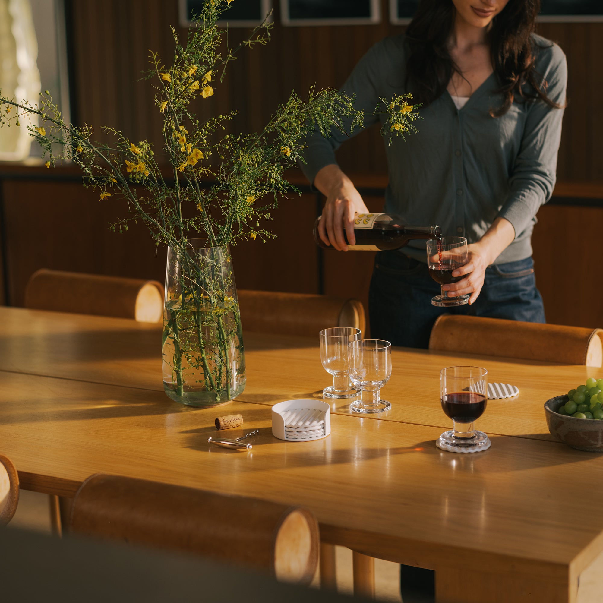 Woman pouring red wine at oak dining table with coaster set, wine glasses, ceramic coaster set, and yellow wildflower centerpiece