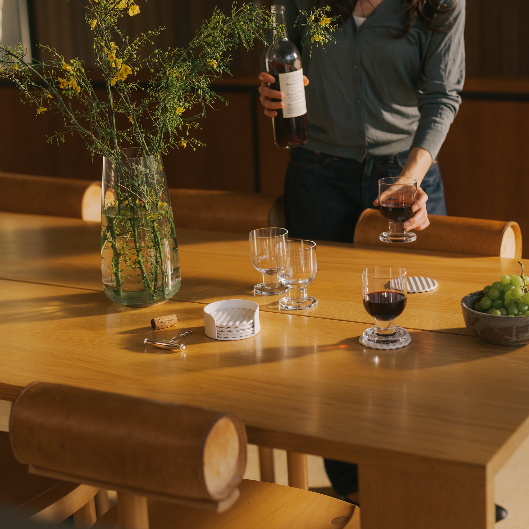 Woman pouring red wine at oak dining table with coaster set, wine glasses, ceramic coaster set, and yellow wildflower centerpiece