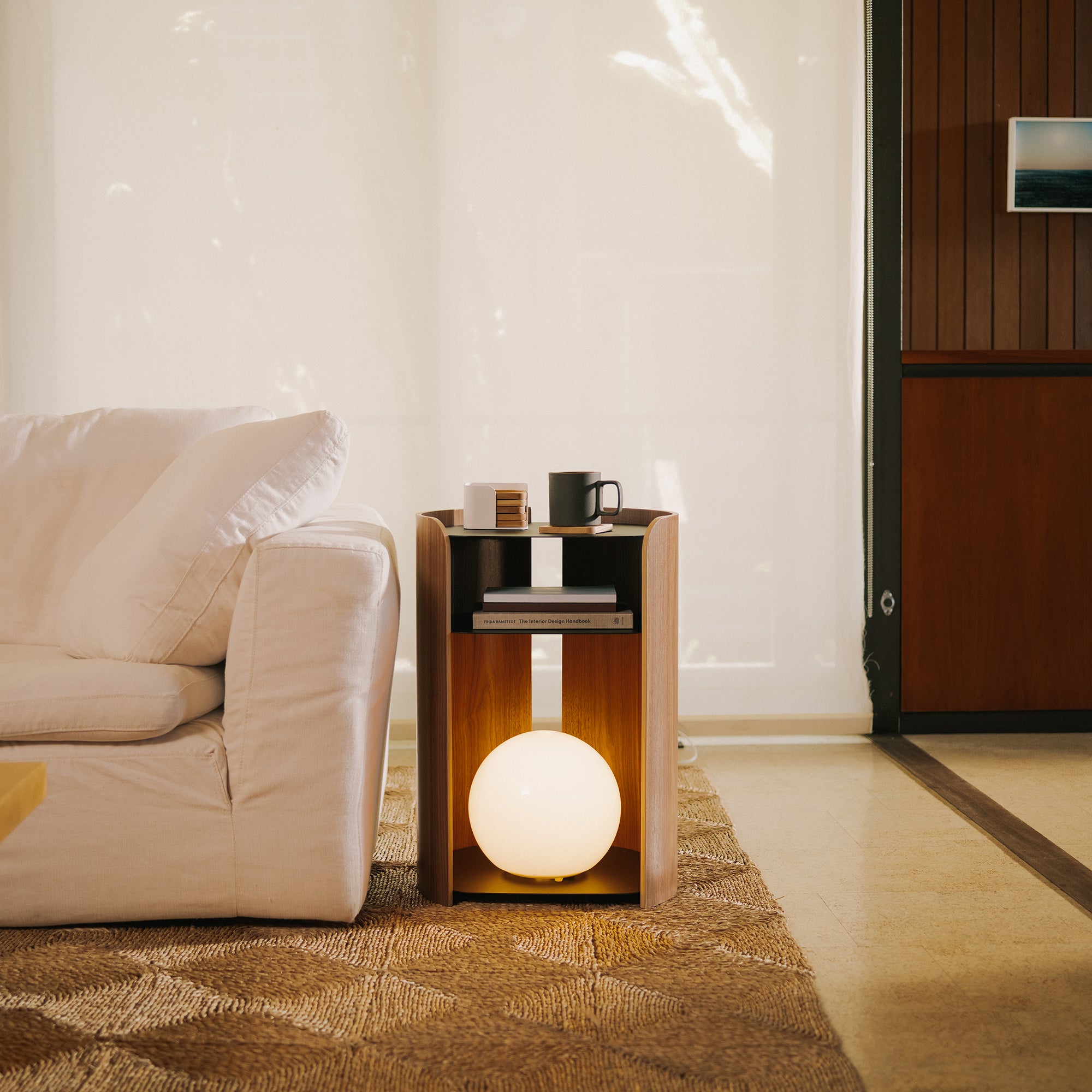 Walnut side table with open shelves, glowing globe lamp, books, and mug beside cream sofa in warm minimalist living room