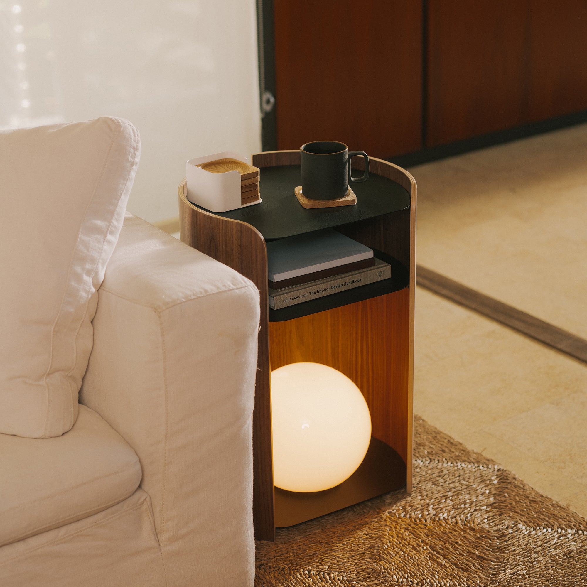 Curved walnut side table with black tray top, stacked books, mug, coasters, and glowing orb lamp beside linen sofa