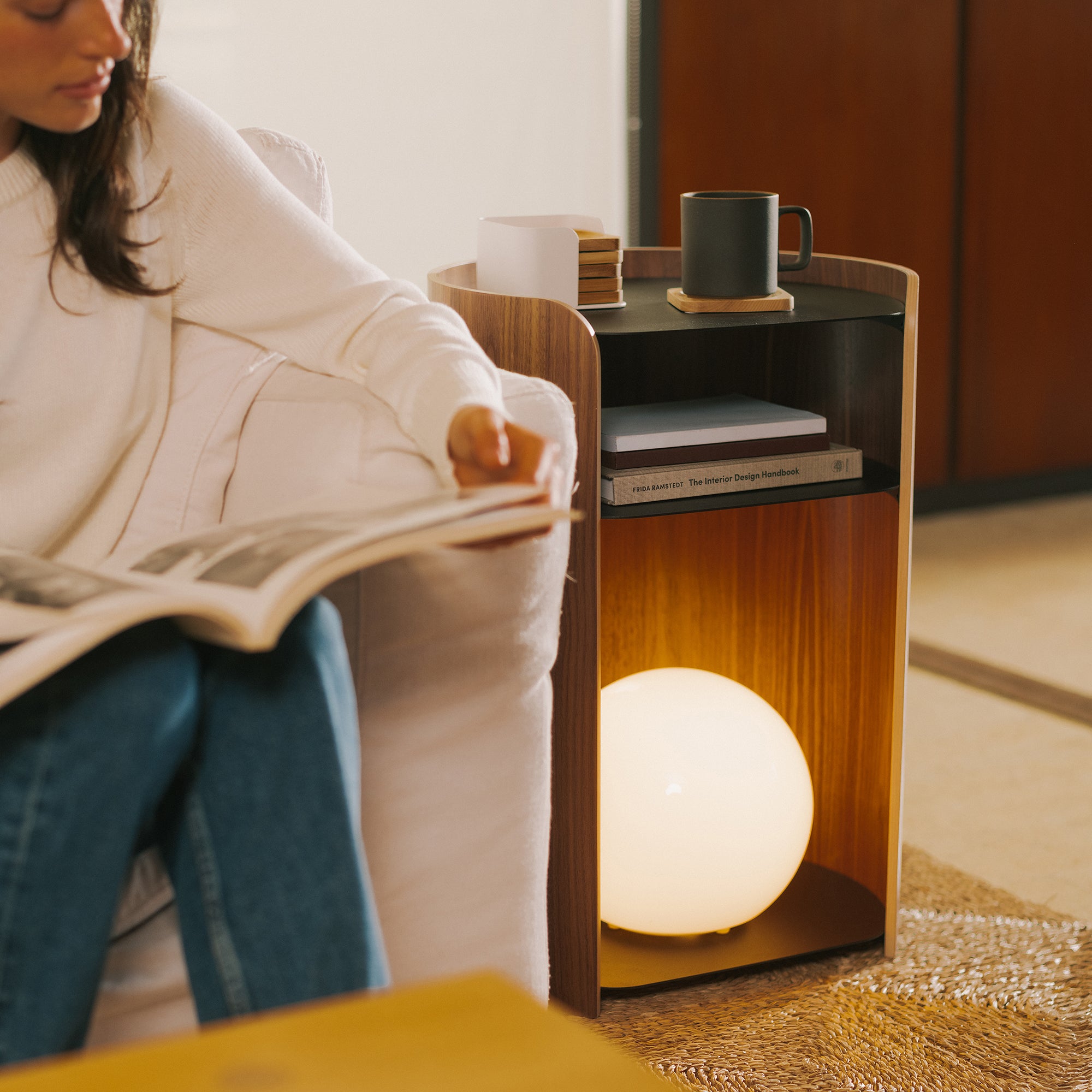 Woman reading magazine on sofa next to walnut side table with glowing orb lamp, books, and mug on black tray top