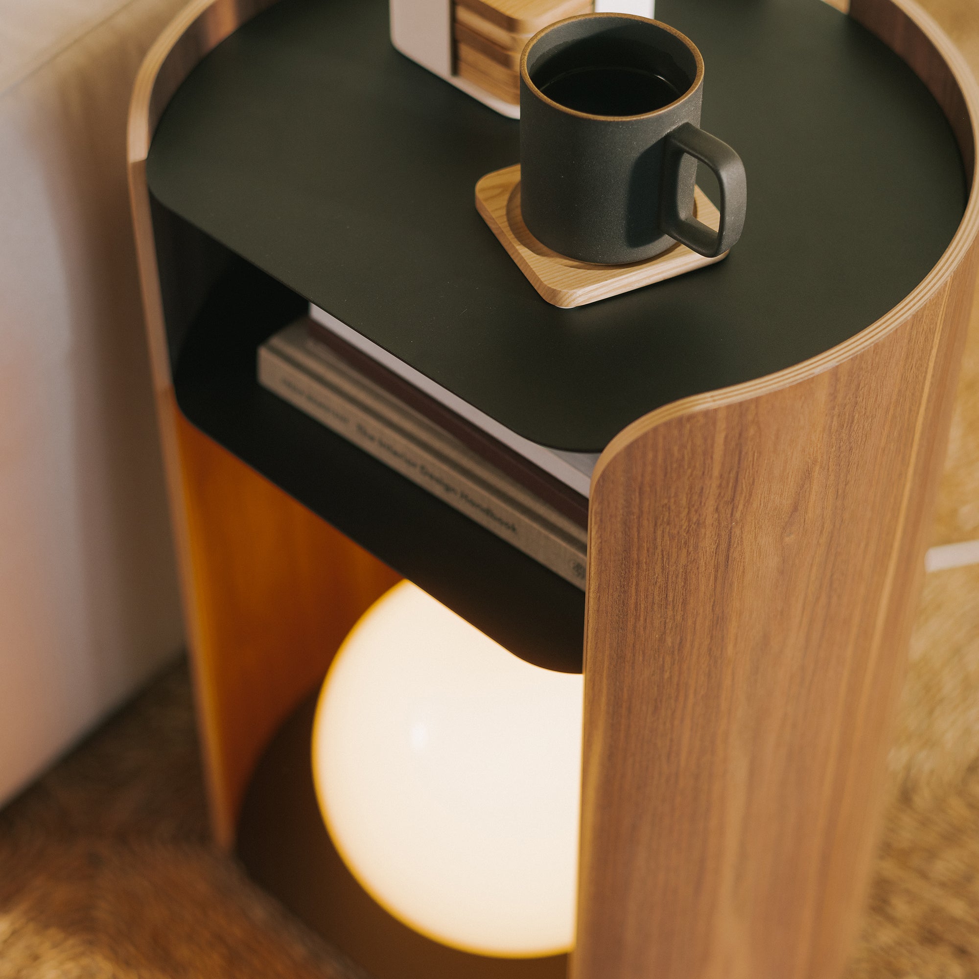 Close-up of curved walnut side table with glowing orb lamp, open book shelf, and matte mug on black tray top