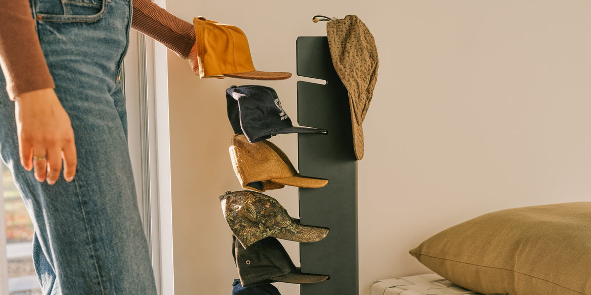 Person grabbing a cap from the Apto Slot Cap Stand in black steel, filled with caps in an entryway beside a bench and cushion
