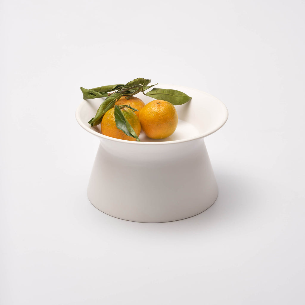 White Apto Flip Fruit Bowl with the wide shallow side up, holding clementines with green leaves, shown on a plain white background