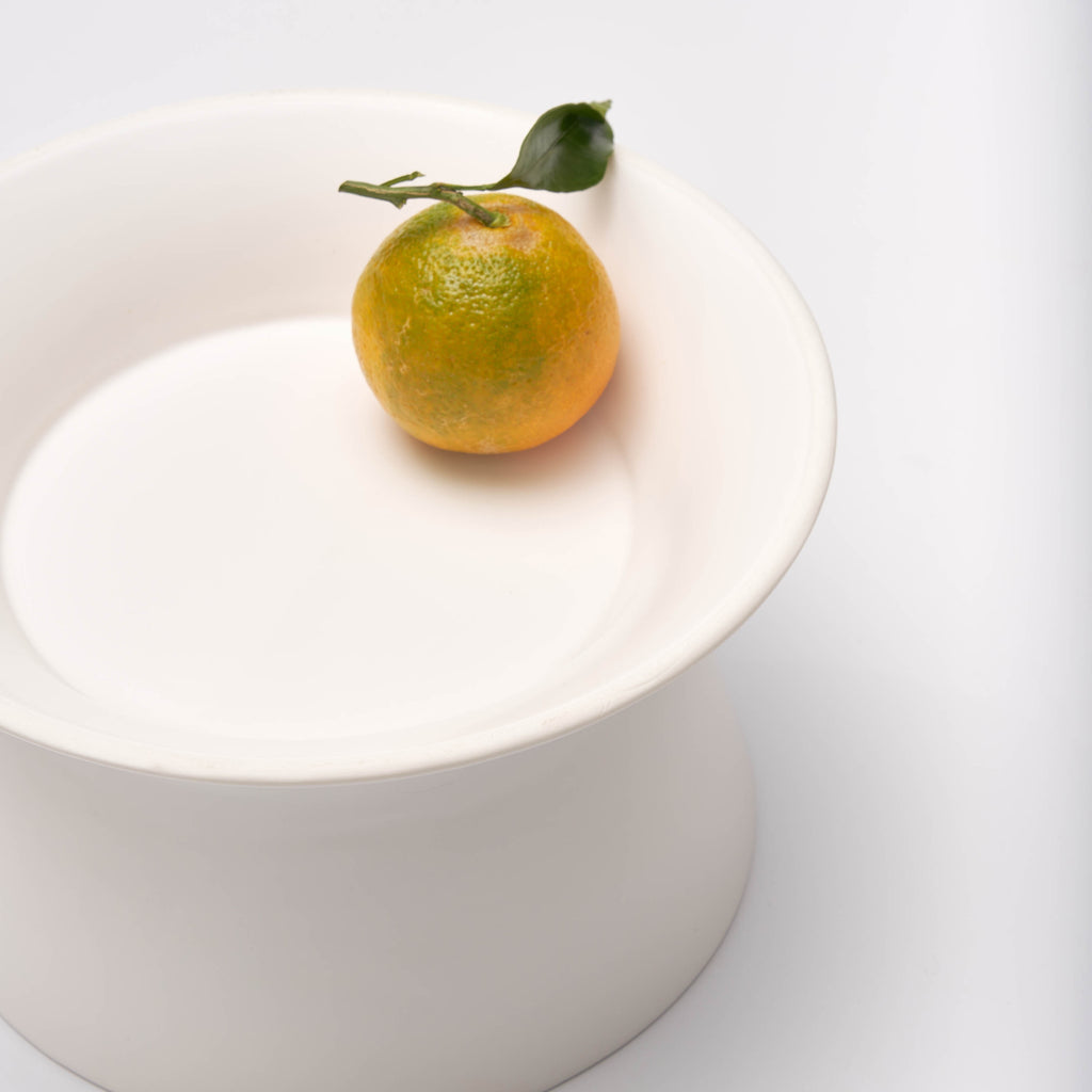 Close-up of a single clementine resting on the wide shallow side of the white Apto Flip Fruit Bowl, shown on a white background