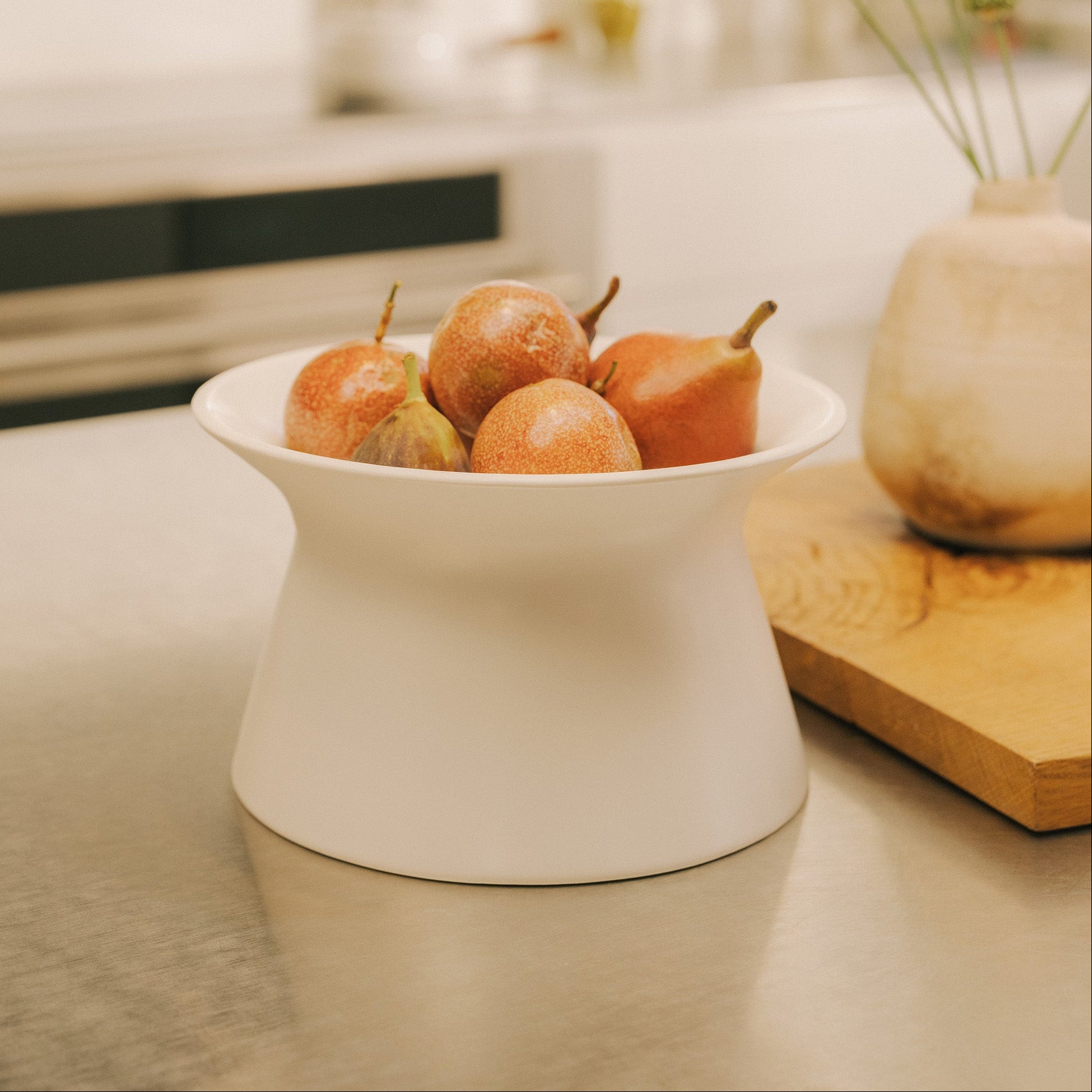 White Apto Flip Fruit Bowl filled with fresh pears on a stainless kitchen counter, next to a wood cutting board and a ceramic vase