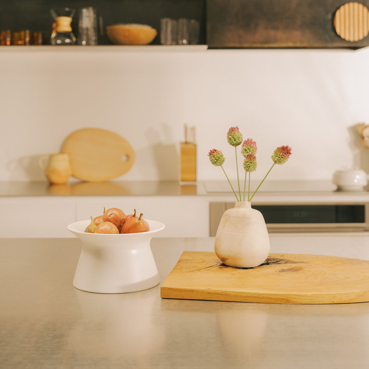 Wide kitchen view of the white Apto Flip Fruit Bowl holding pears on the counter, beside a wood board and a vase of fresh flowers
