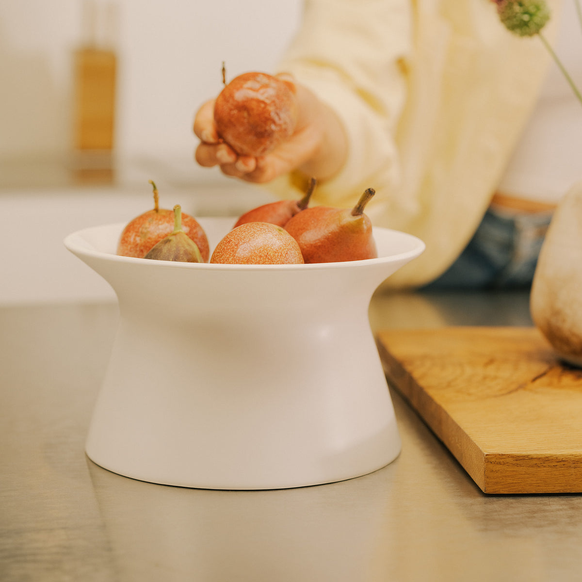 Hand picking a pear from the white Apto Flip Fruit Bowl on a kitchen counter, with a wood cutting board and vase in the background