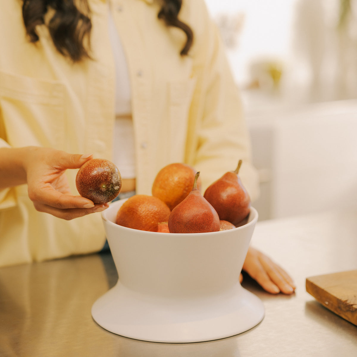 Woman reaching for a pear from the white Apto Flip Fruit Bowl filled with fresh fruit on a stainless steel kitchen counter at home