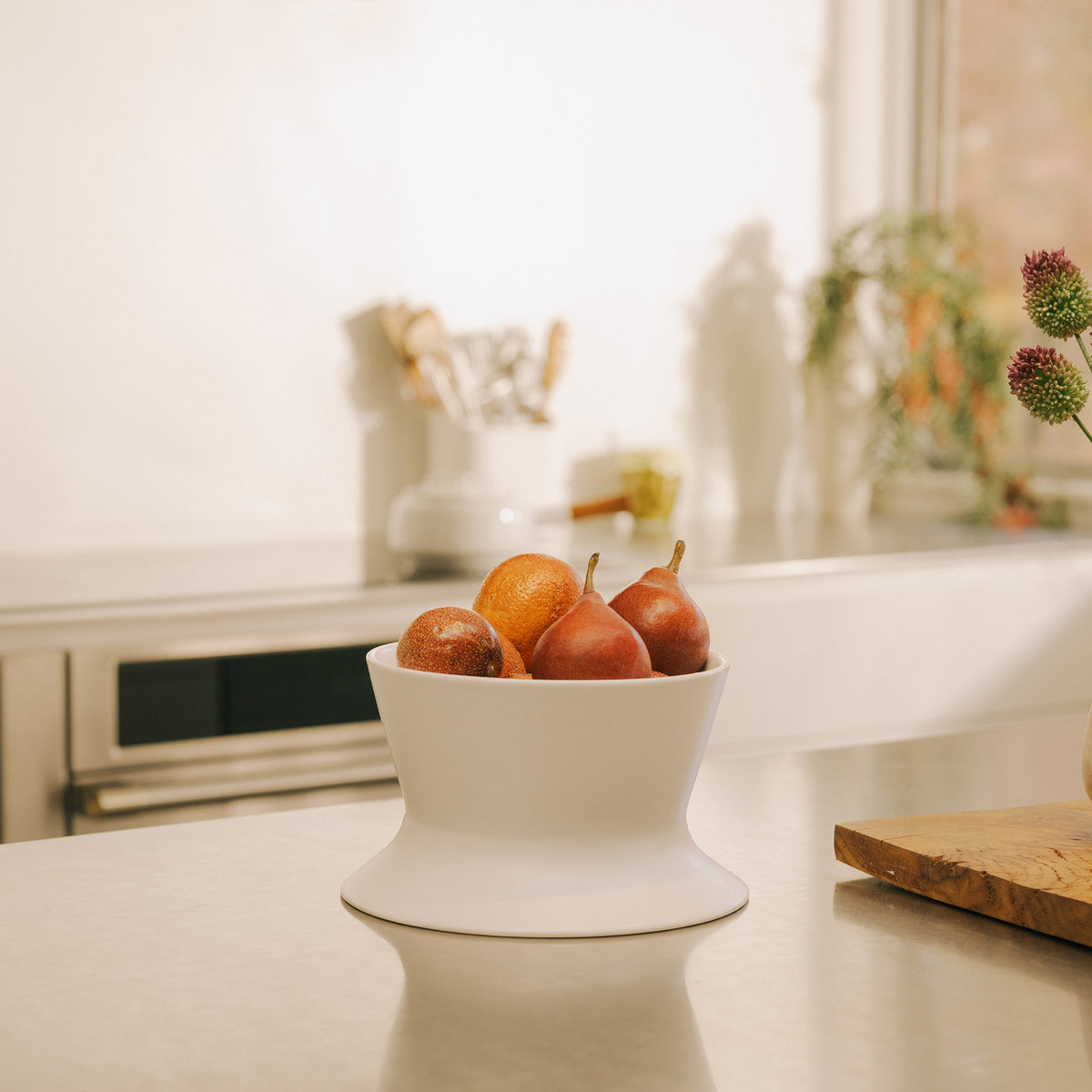 White Apto Flip Fruit Bowl holding fresh pears on a stainless steel counter in a bright kitchen with plants and a window behind it