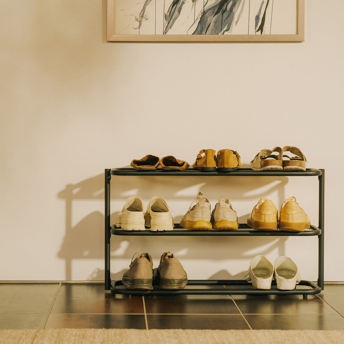 Front view of the Apto Obround Shoe Rack Short in black steel filled with sneakers, loafers and sandals in a sunny entryway with framed art