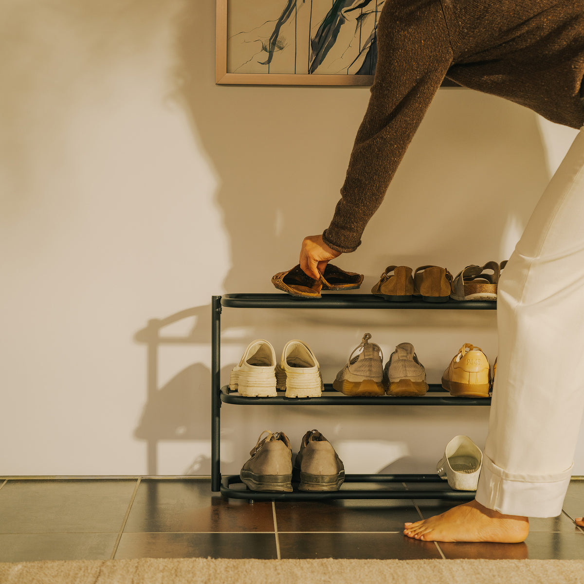 Person placing a pair of shoes on the top tier of the Apto Obround Shoe Rack Short in black steel, filled with shoes in a sunlit entryway
