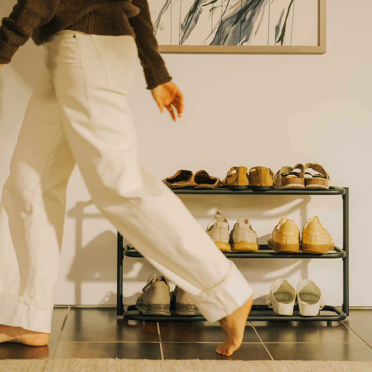 Person walking barefoot past the Apto Obround Shoe Rack Short in black steel, filled with shoes on three tiers in a sunny entryway
