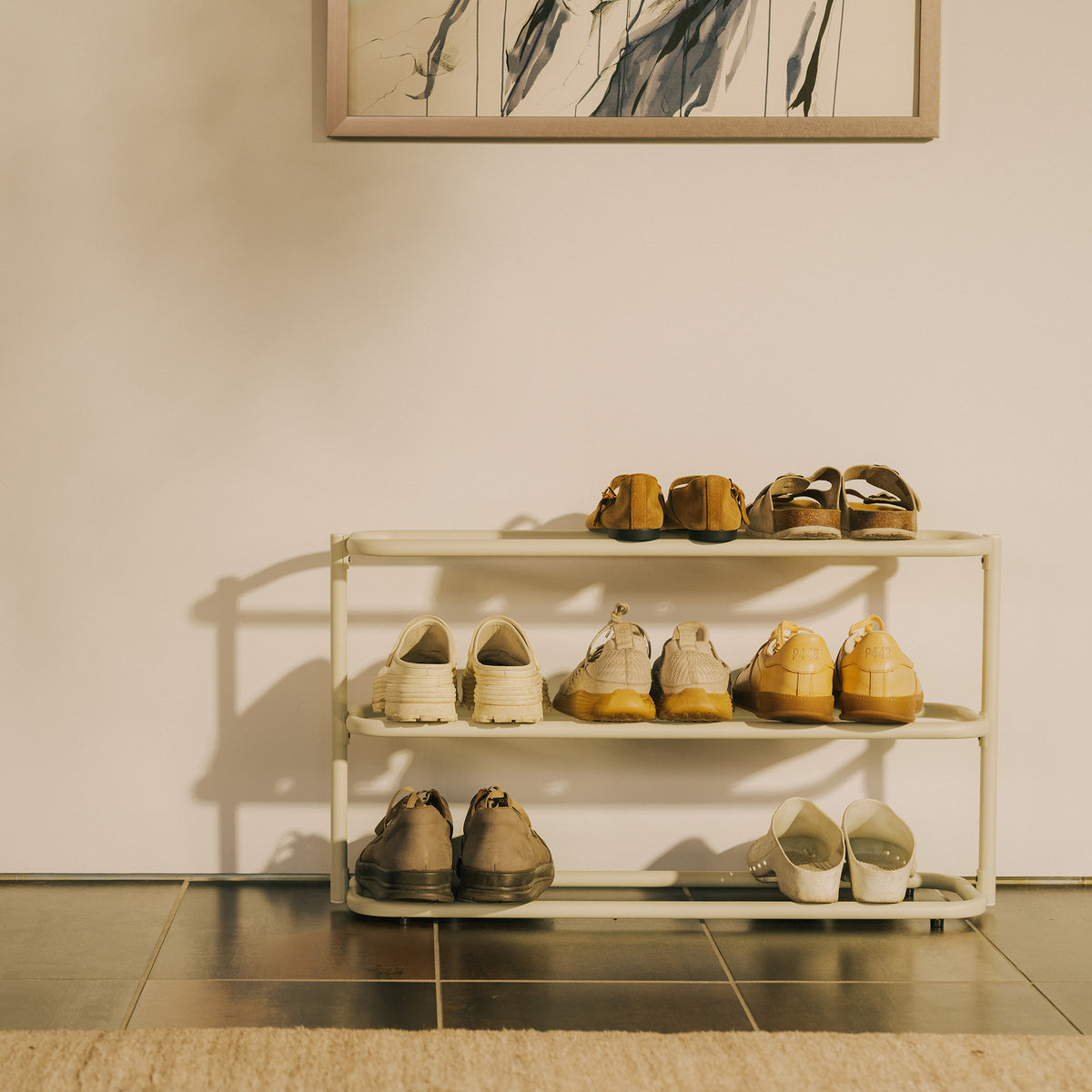 Front view of the Apto Obround Shoe Rack Short in ivory steel holding shoes on three tiers in an entryway with framed art above
