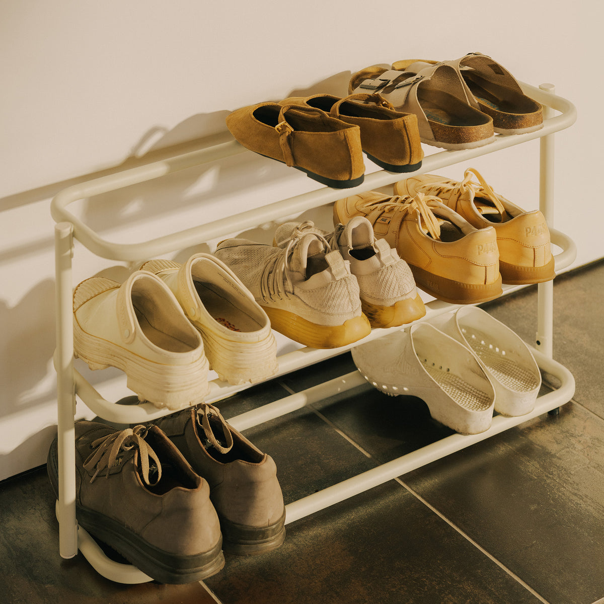 Close-up of the Apto Obround Shoe Rack Short in ivory steel holding loafers, sneakers and clogs on three tiers on a tile floor