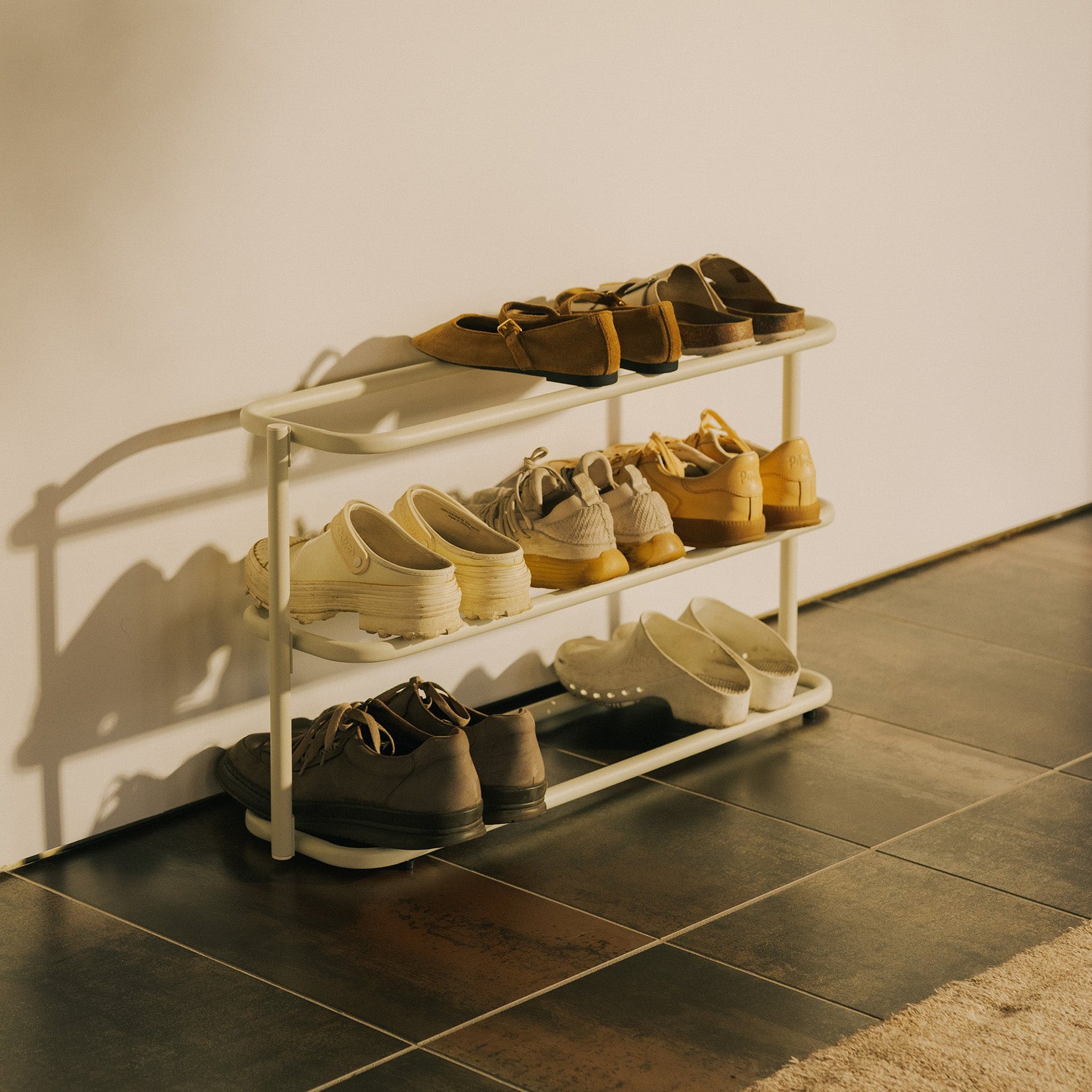 Angled view of the Apto Obround Shoe Rack Short in ivory steel filled with shoes on three tiers in a sunny entryway with tile floor