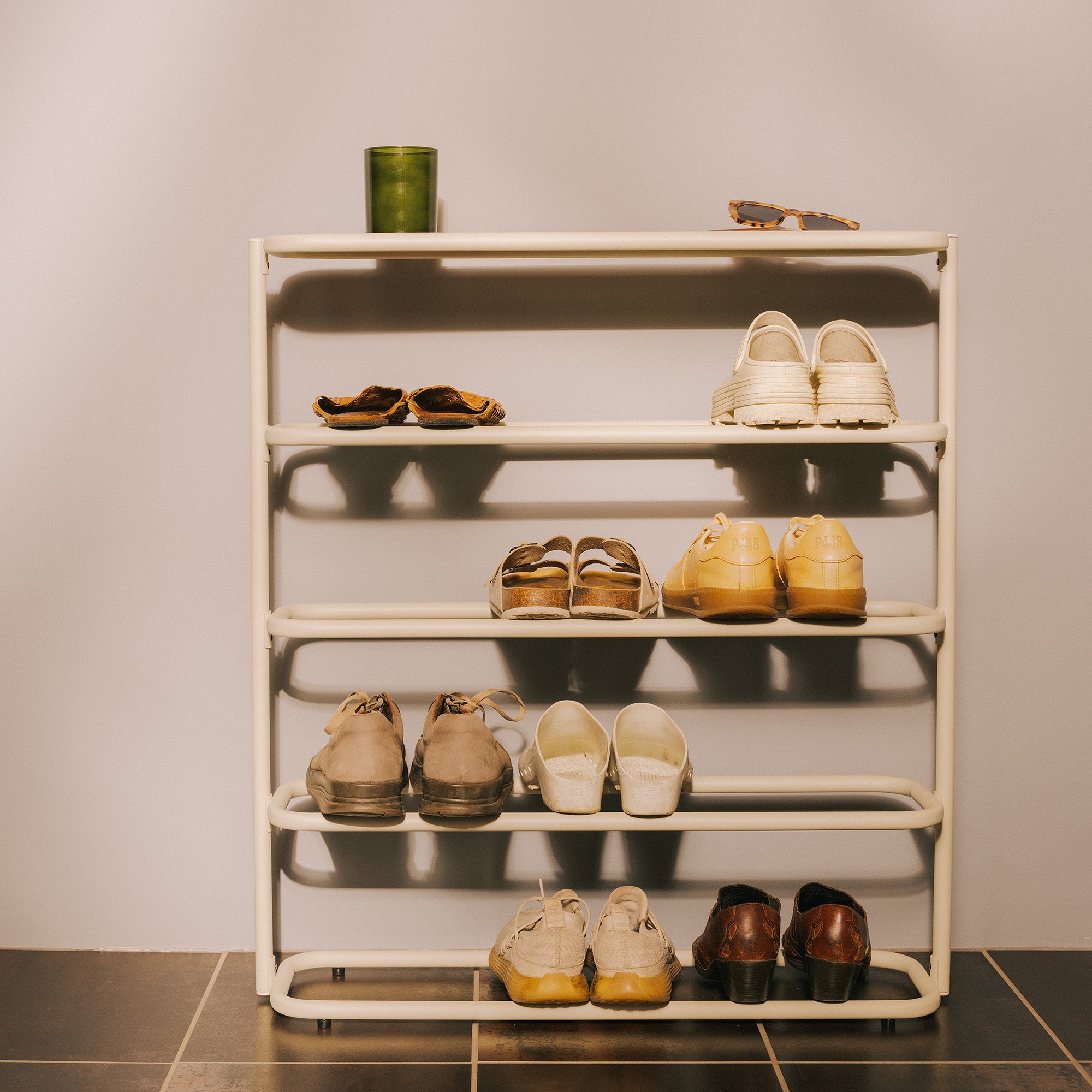 Ivory 4-tier metal shoe rack with tabletop against a white wall, holding 8 pairs of shoes, topped with sunglasses and candle.