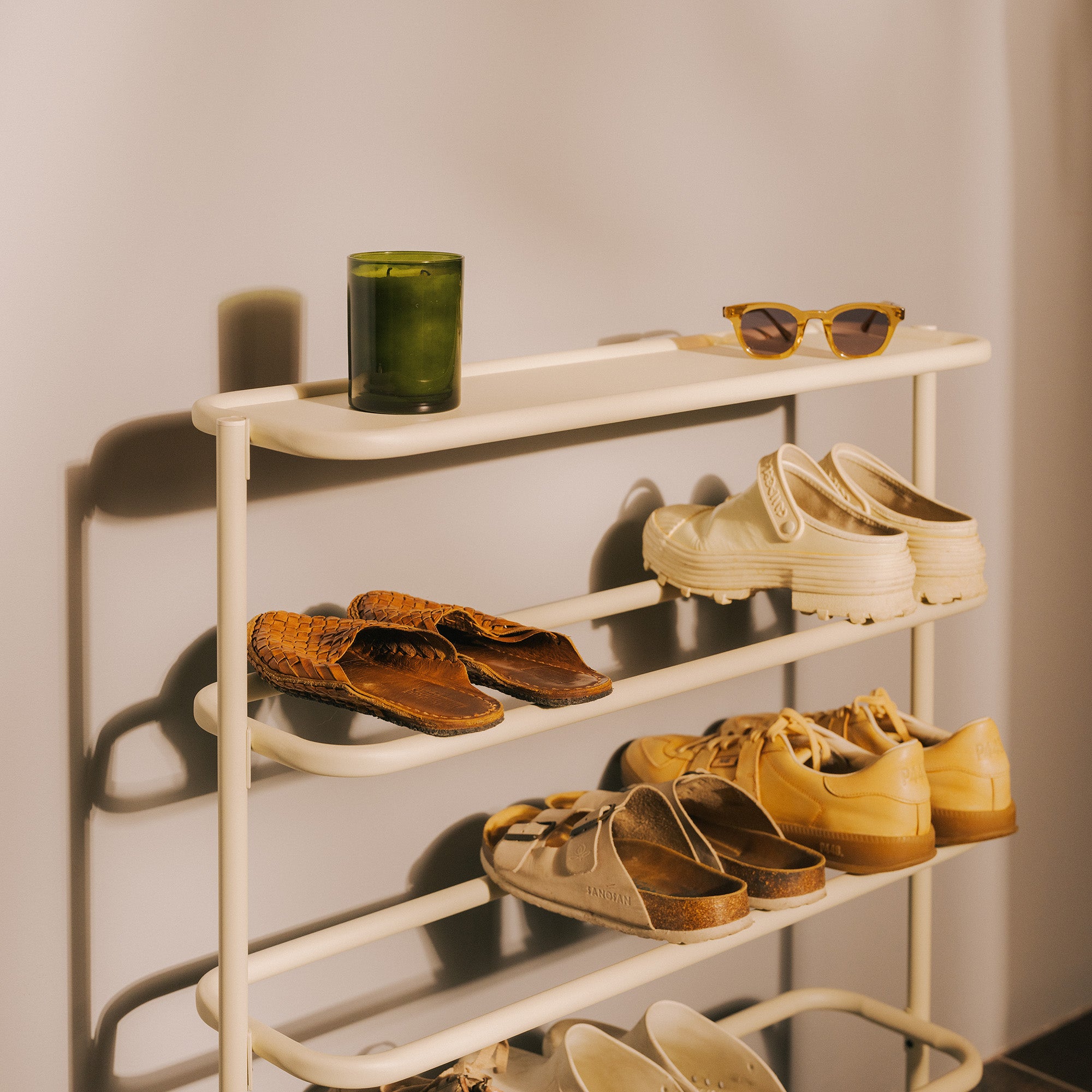 Ivory metal entryway shoe rack with flat top shelf holding sunglasses, candle, filled with shoes