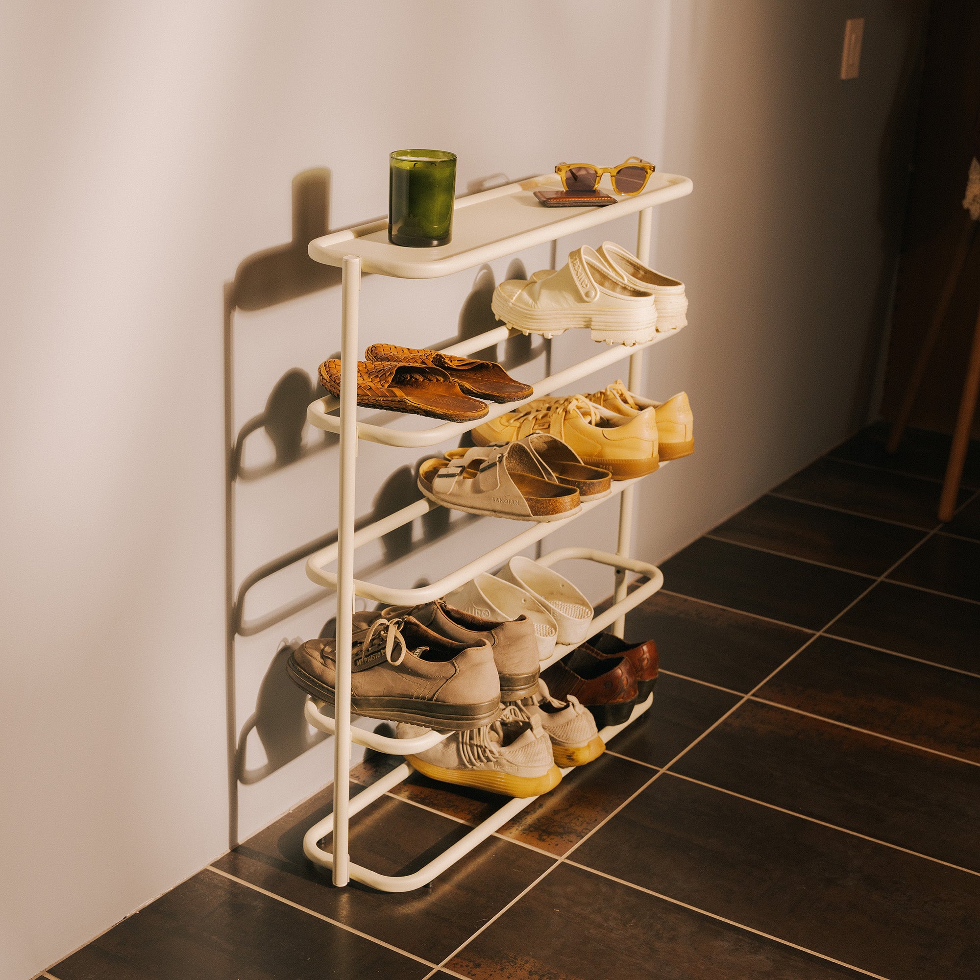 Ivory metal 4-tier shoe rack organizer for entryways, styled with sunglasses and candle against white wall