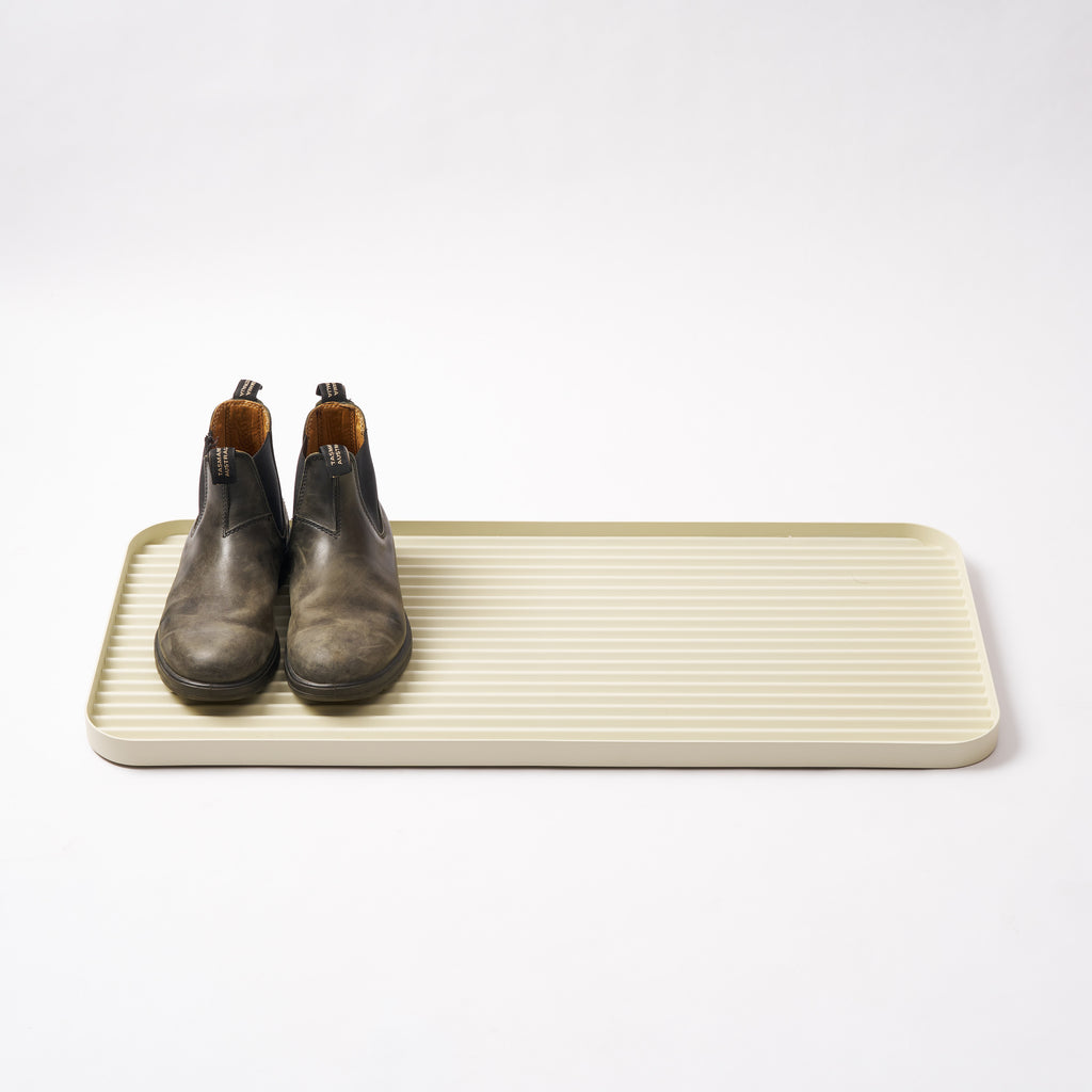 Cream steel boot tray with ridged bottom holding a pair of boots with a white backdrop. Designed by Apto. 
