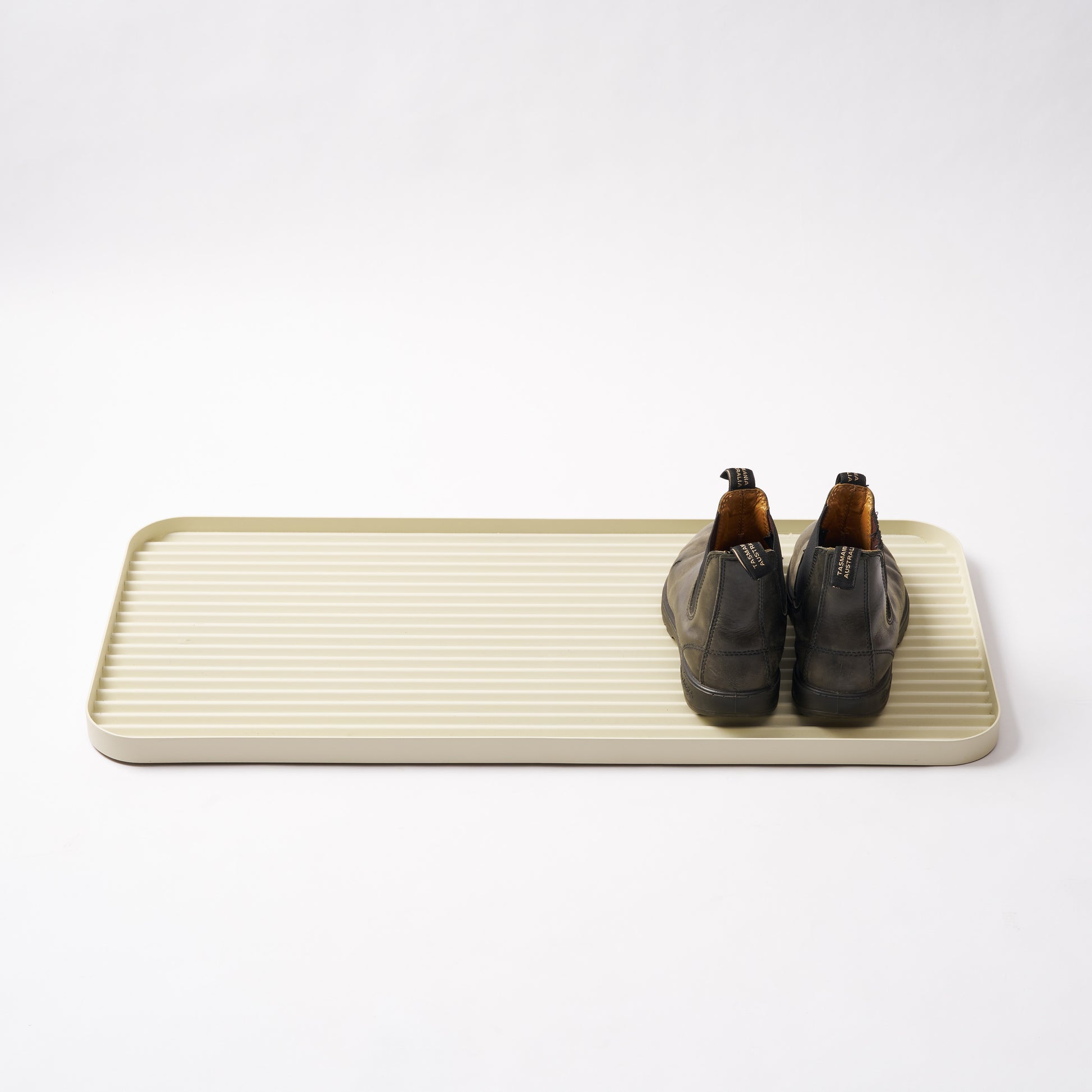 Ivory steel boot tray with ridged bottom holding a pair of boots with a white backdrop. Designed by Apto. 