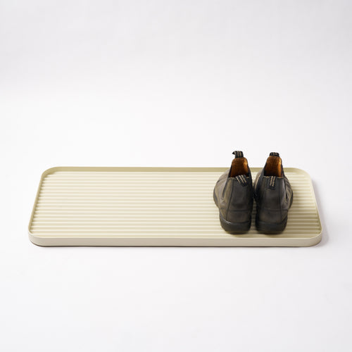 Ivory steel boot tray with ridged bottom holding a pair of boots with a white backdrop. Designed by Apto. 