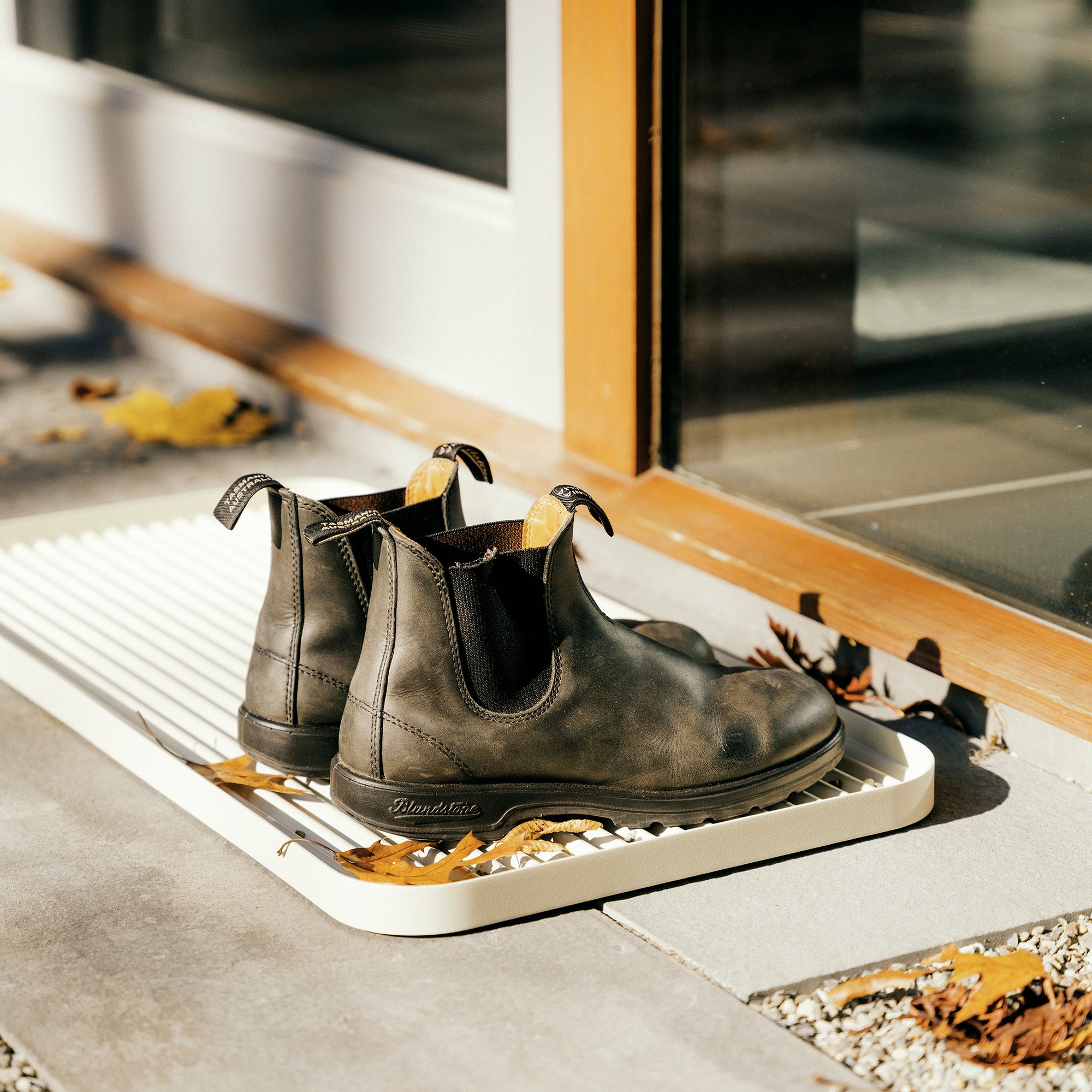 White steel boot tray with ridged bottom holding a pair of Blundstones and fall leaves outside a modern home.