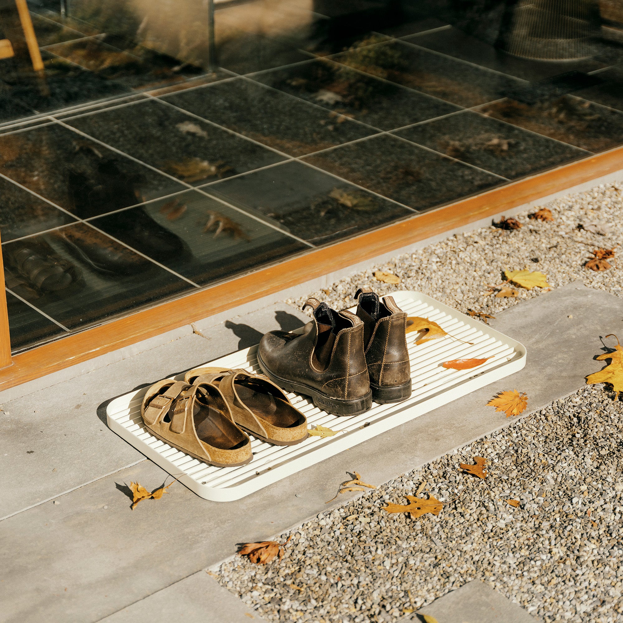 White steel boot tray with ridged bottom on a concrete patio holding a pair of Birkenstocks and Blundstones and fall leaves outside a modern home.