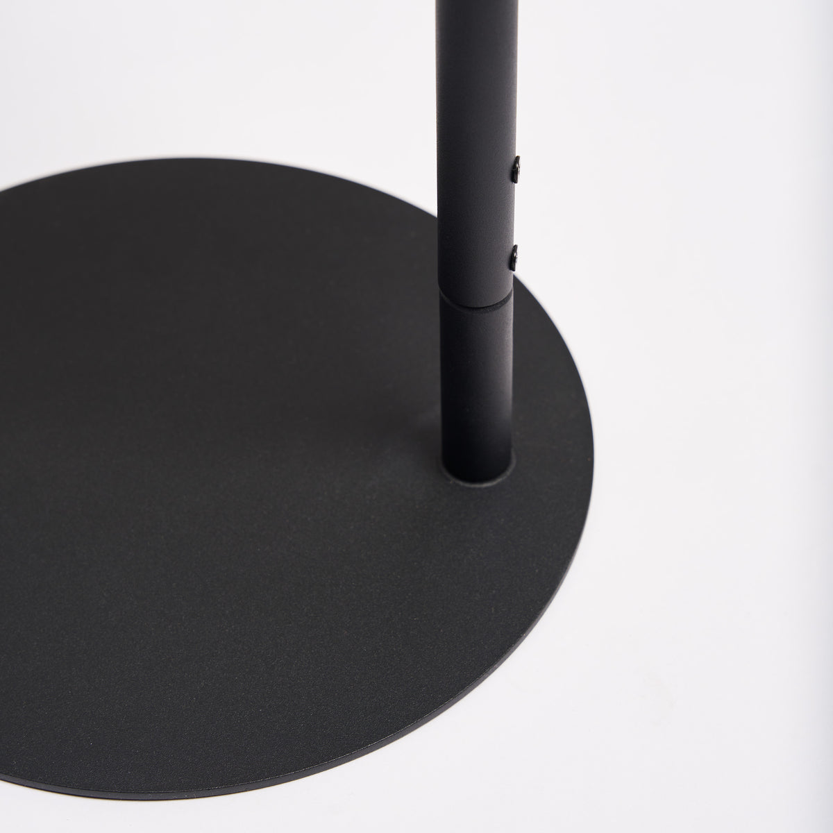 Close-up of the round base on the Apto Slot Cap Stand in black steel, showing the slim pole and the flat, sturdy footprint, on a white background