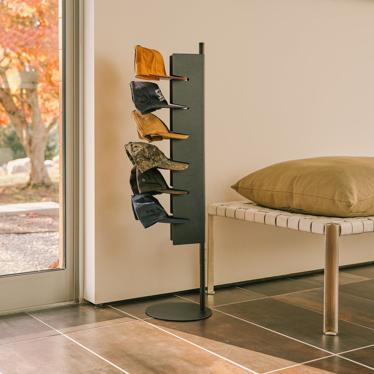 Apto Slot Cap Stand in black steel holding six baseball caps by a sunny window in an entryway, next to a bench with a cushion