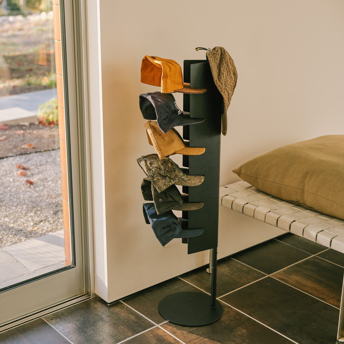 Apto Slot Cap Stand in black steel holding baseball caps and a bucket hat in a bright entryway, with a bench and window beside it