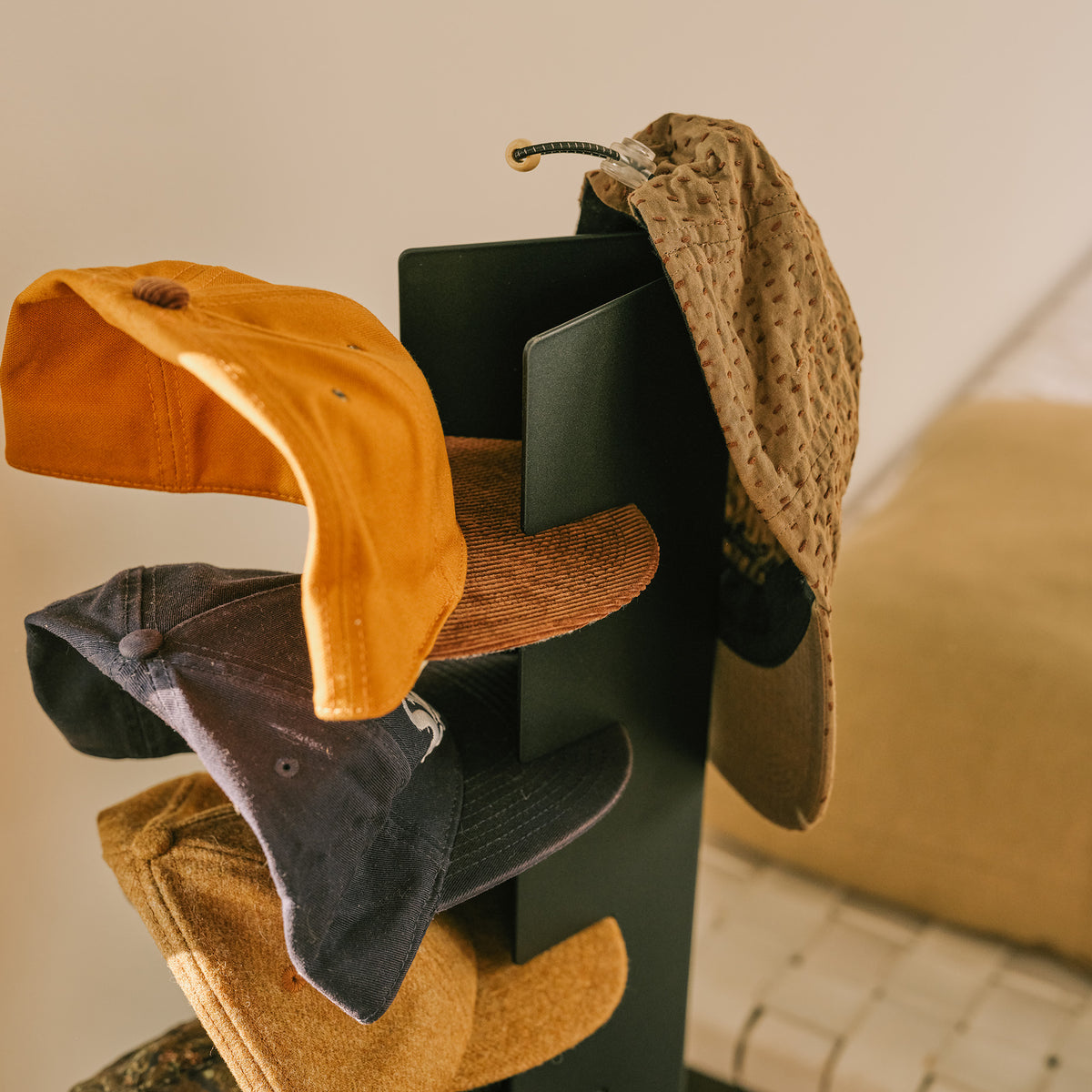 Close-up of baseball caps hooked into the slots of the Apto Slot Cap Stand in black steel, with a knit bucket hat hanging on the top