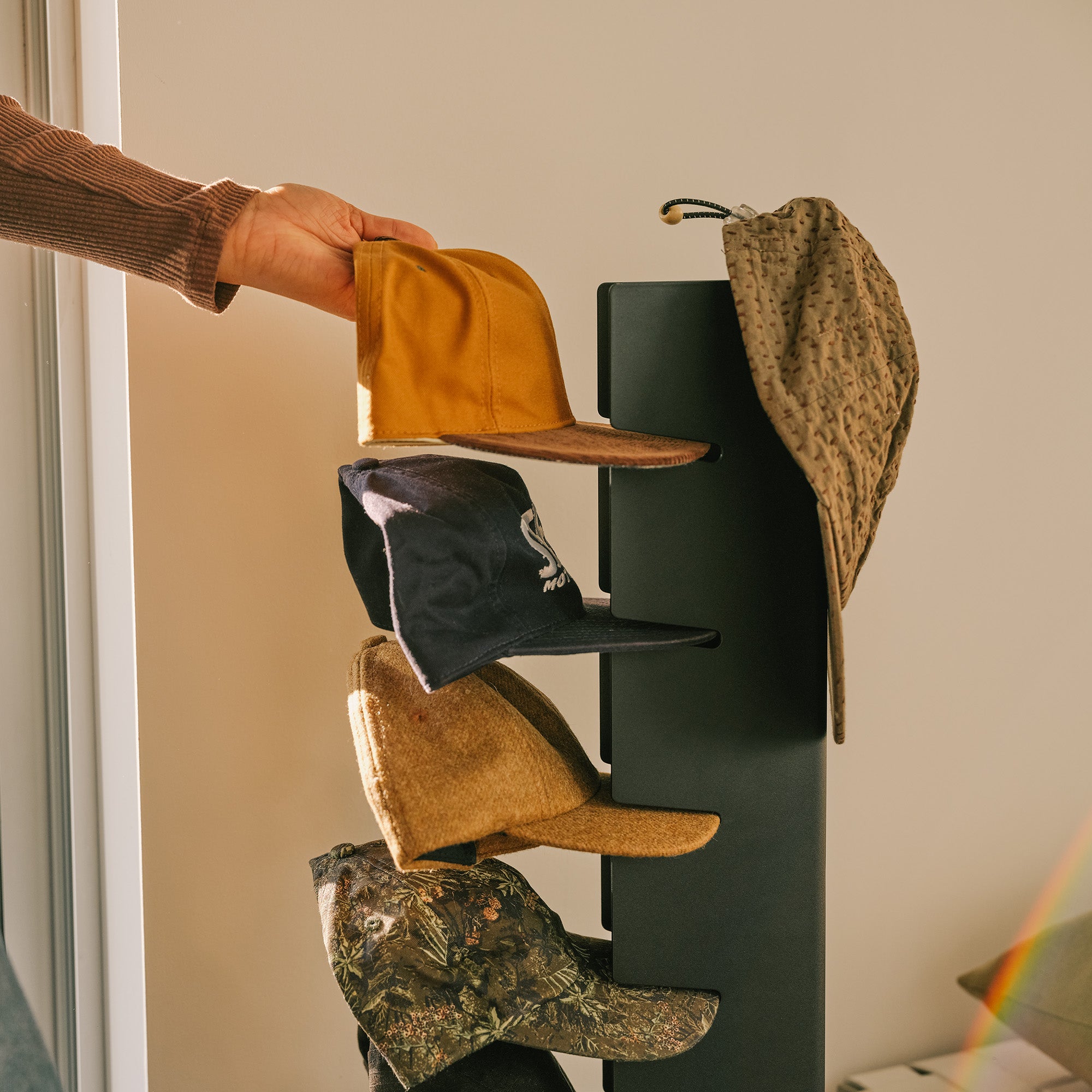 Hand hanging a baseball cap on the Apto Slot Cap Stand in black steel, which holds a neat stack of caps and a bucket hat by the door