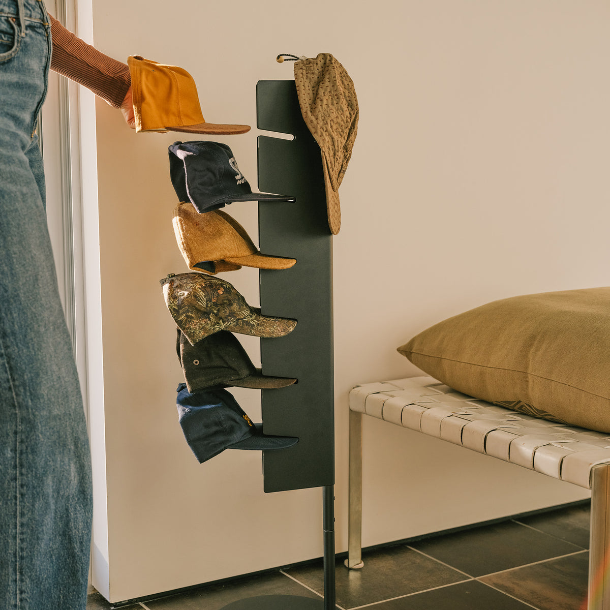 Person grabbing a cap from the Apto Slot Cap Stand in black steel, filled with caps in an entryway beside a bench and cushion