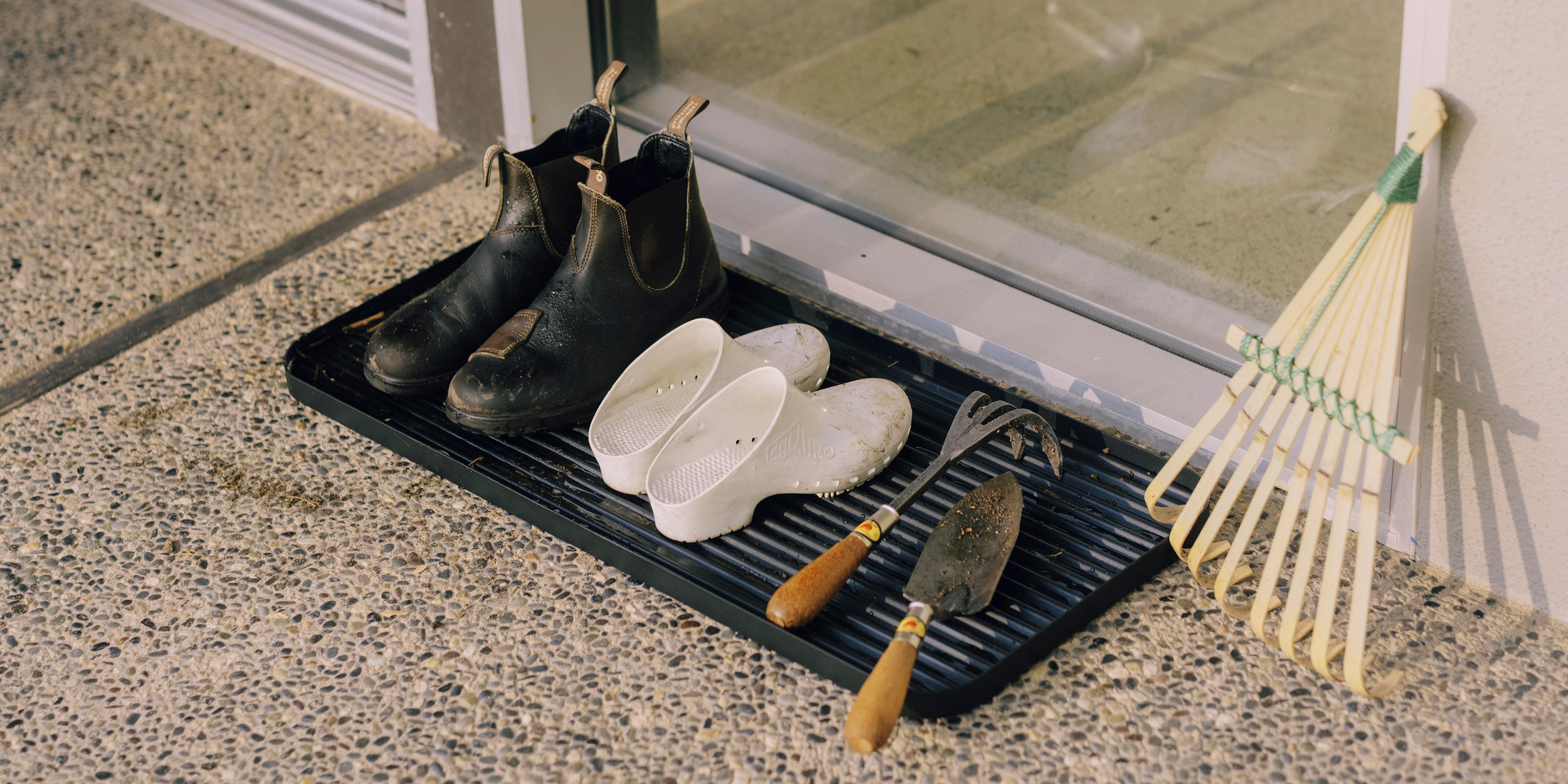 Black steel boot tray from Apto outside a front door holding two pairs of shoes and gardening supplies.