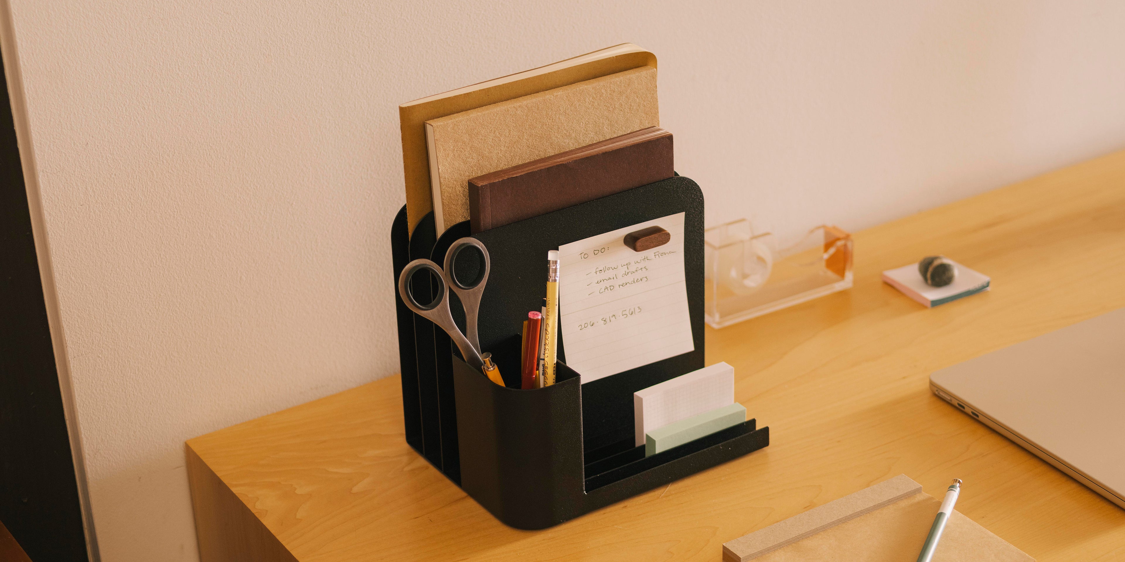 Black steel desk caddy with using slots for pens, files, and sticky notes designed by Apto on a modern wood desk with a computer and other design tools. 