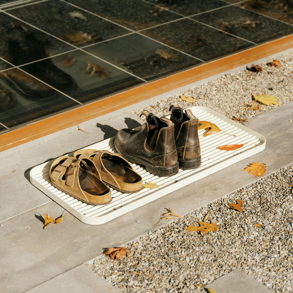 White steel boot tray with ridged bottom on a concrete patio holding a pair of Birkenstocks and Blundstones and fall leaves outside a modern home.